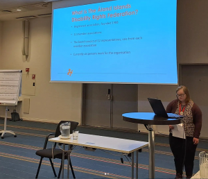 Kvinna med glasögon och halvlångt hår står framför powerpoint-presentation där det står "We are the Åland Disability federation"