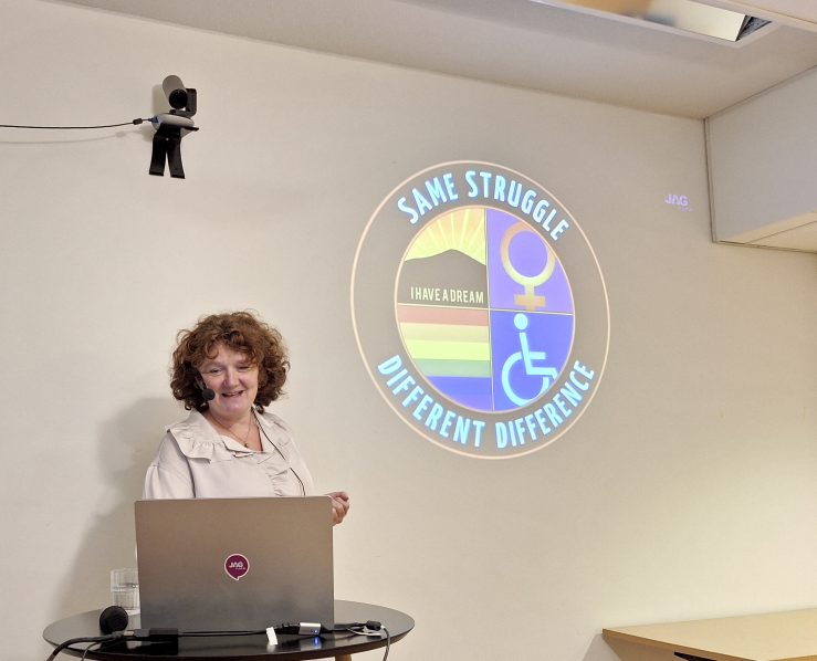 Kvinna med lockigt hår som står framför en dator. På skärm bakom texten: Same struggle different difference och bild på rullstolsmärke, prideflagga mm.