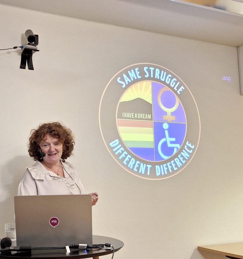 Kvinna med lockigt hår som står framför en dator. På skärm bakom texten: Same struggle different difference och bild på rullstolsmärke, prideflagga mm.