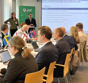 Foto visar 2 personer längst fram som håller presentation, framför syns åhörare vid konferensbord samt nordiska flaggor