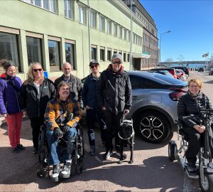 Foto visar grupp av personer framför bil och grön byggnad. Två i grupen har rullstol, en person har med sig ledarhund.