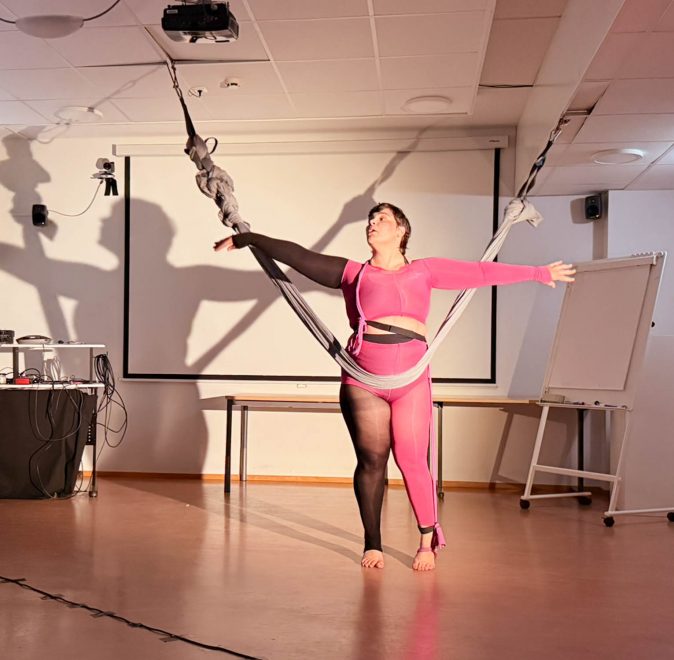 Foto visar kvinna i orange och svart kroppsstrumpa som dansar med ett grått tygstycke som hänger från taket