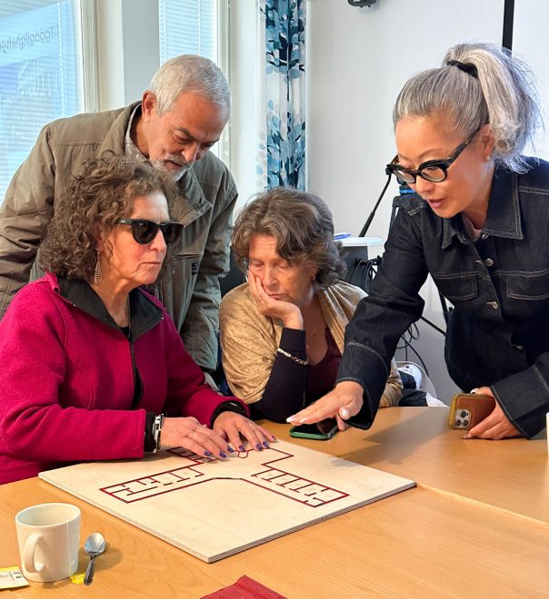 Foto visar fyra personer framför taktil träskylt föreställande ritning av byggnad, en av personerna känner på den.