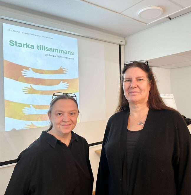 Foto visar två kvinnor i svarta kläder framför skärm som visar bild av händer