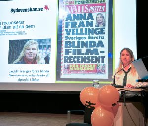 Foto visar kvinna på scen framför skärm med löpsedel