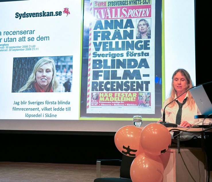 foto visar kvinna på scen framför skämr med löpsedel