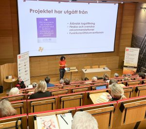 Foto av publik bakifrån, framme vid stor skärm en föreläsare