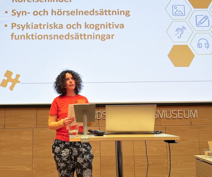 Foto av kvinna som föreläser framför skärm, hon har orange tröja