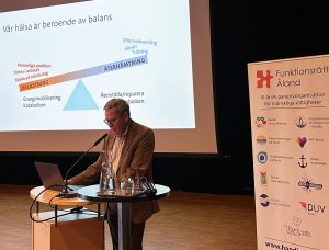 Foto visar man framför mikrofon med presentation i bakgrunden